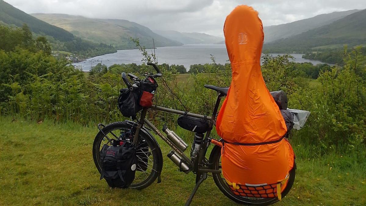 A black bike with a large orange bag strapped to the back with mountains and a lake in the background.
