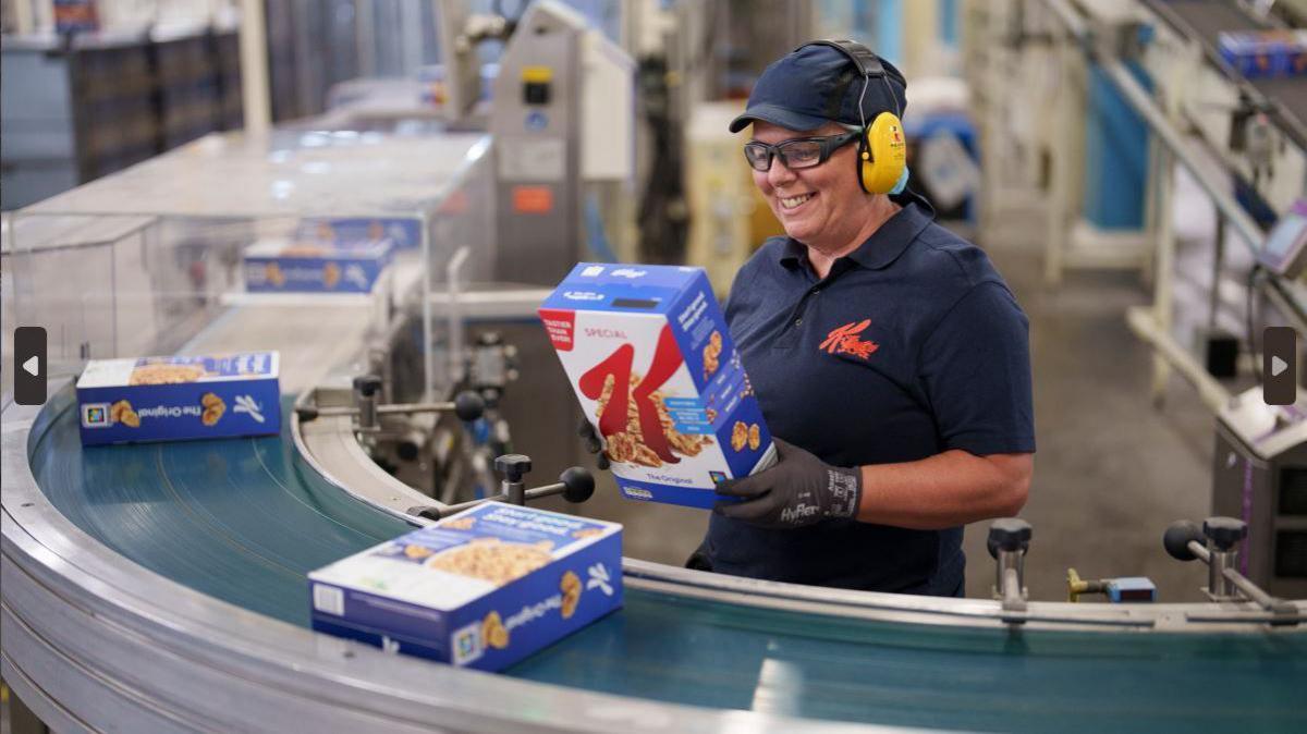 A woman in a navy Kellogg's top with a navy hat and yellow ear protectors. She is stood at the conveyor belt and she is picking up a Kellogg's box which is blue and there are two other boxes are on the belt.