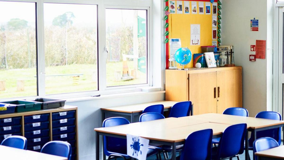 A classroom with a number of tables with blue chairs. There is a large window on one of the walls.