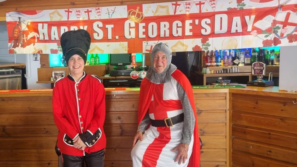 A woman in red Beefeater outfit with her hands clasped and a man in a medieval knights uniform with an English flag and belt. A bar setting and wooden table is behind them both with a flag which says 'Happy St George's Day'