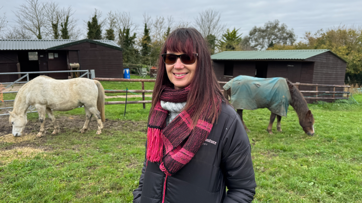 A woman with long dark hair, dark glasses, a black coat and pink and black scarf stands in front of two horses grazing in a field. There are wooden stables in the background.