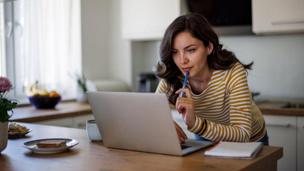 A generic photo of a woman in a striped brown and white top looking at a laptop that is on a surface. Cupboards are blurred behind her.