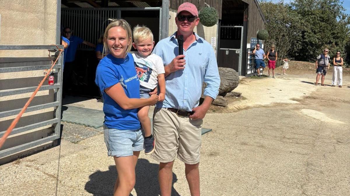 Rachel, Thomas and Joe stood outside their horses stables on a sunny day. There are a few people standing in the background.