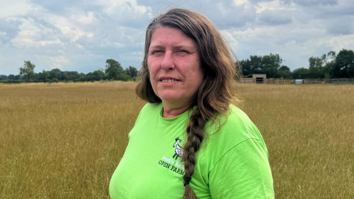 A woman stands alone in a wide field of hay beneath a cloud-filled sky. She wears a bright green T-shirt and has long, braided hair. Trees line the distant horizon.