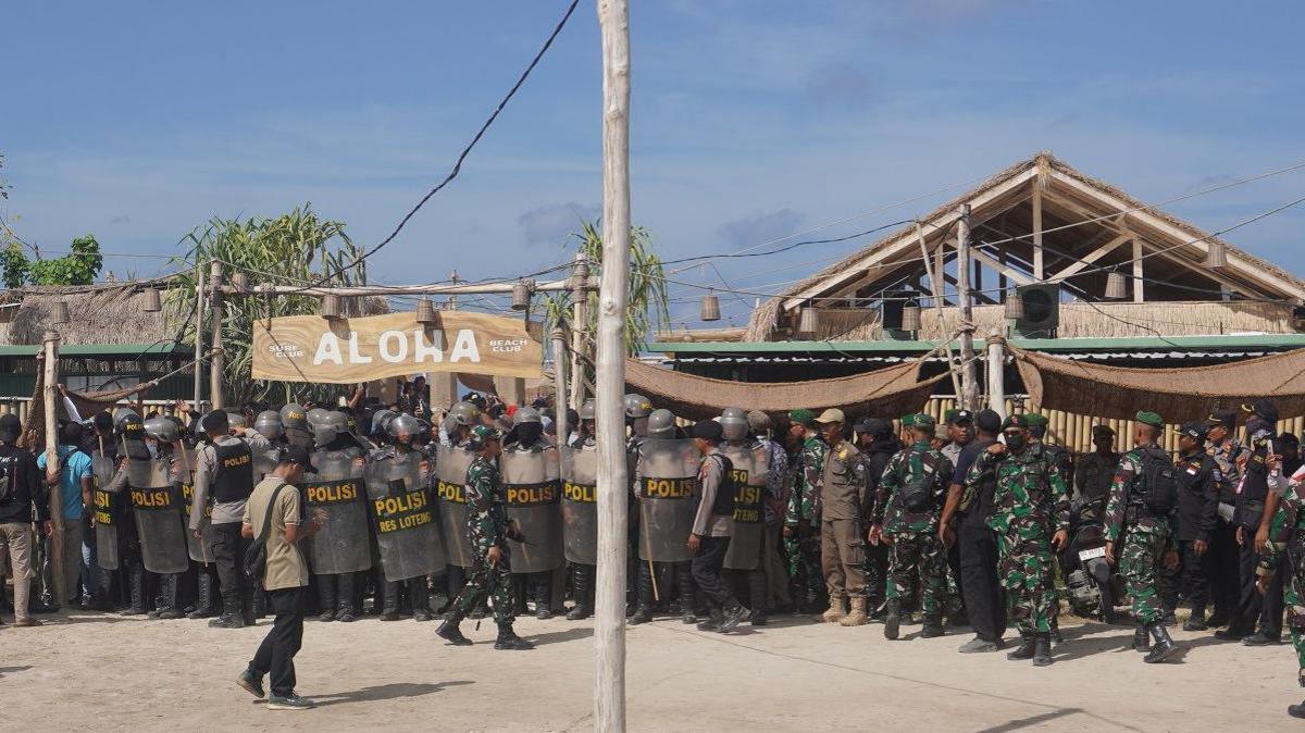 Police with helmets and riot shields, as well as men in green camouflge uniform, crowd in front of a hut that has a large wooden sign that reads "Aloha".
