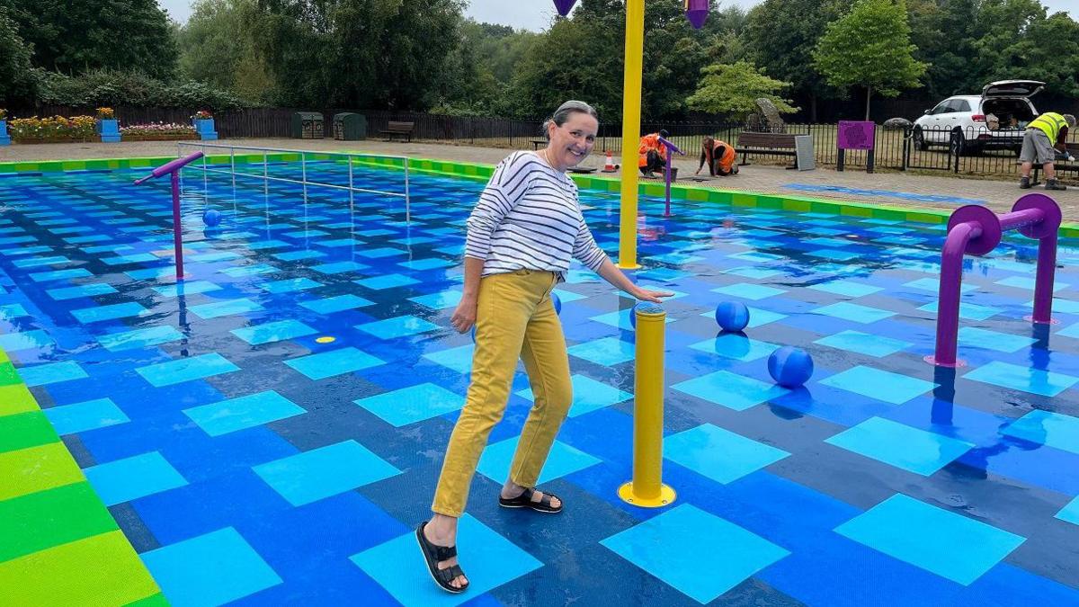 A woman wearing a stripy black and white top, yellow trousers and black sandals rests her hand above a water feature in a splash park