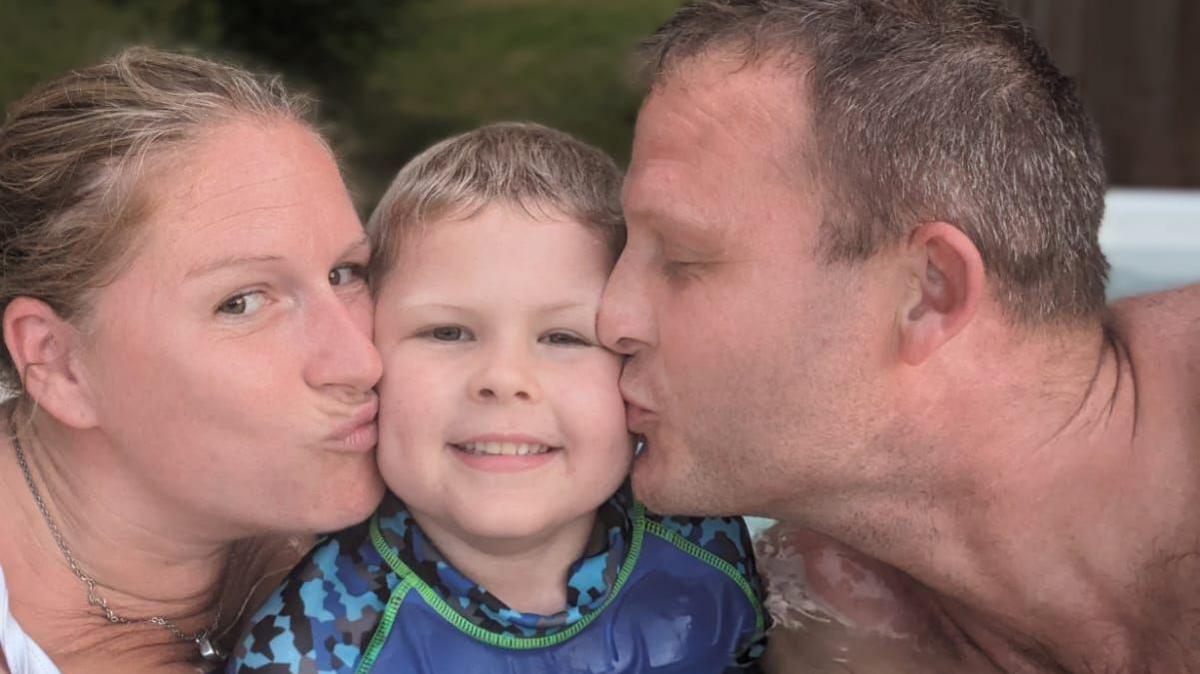 A woman and a man are on either side of Bodhi, kissing each cheek. Bodhi is smiling and looking straight at the camera. 