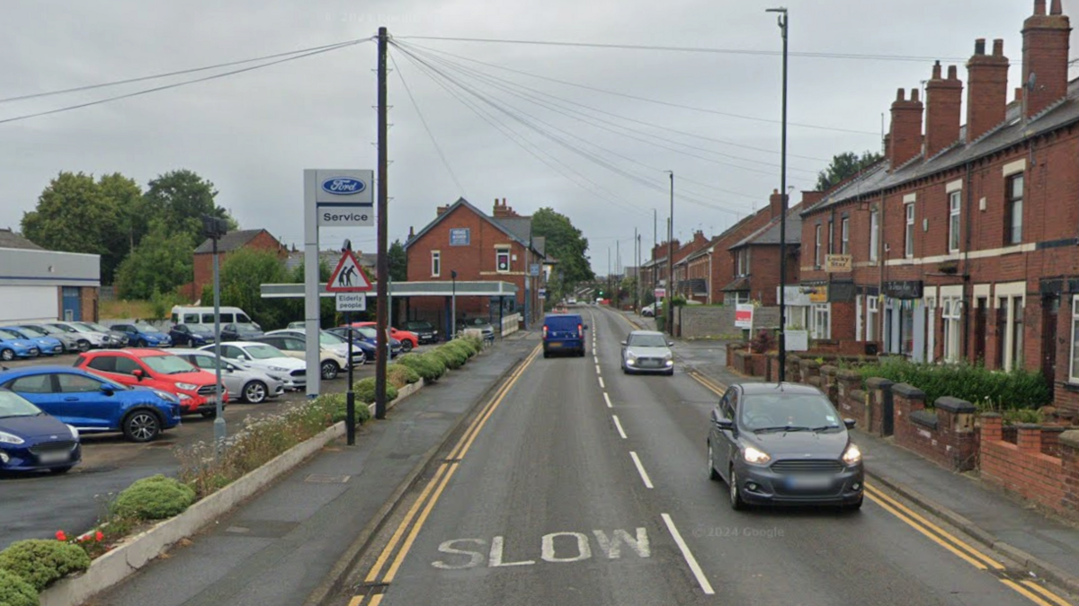 A two-lane urban road with the word “SLOW” painted on the asphalt. Several cars are driving along the street, and parked vehicles line both sides. On the left, there is a Ford service centre. On the right, a row of red-brick terraced houses with chimneys and small front gardens.