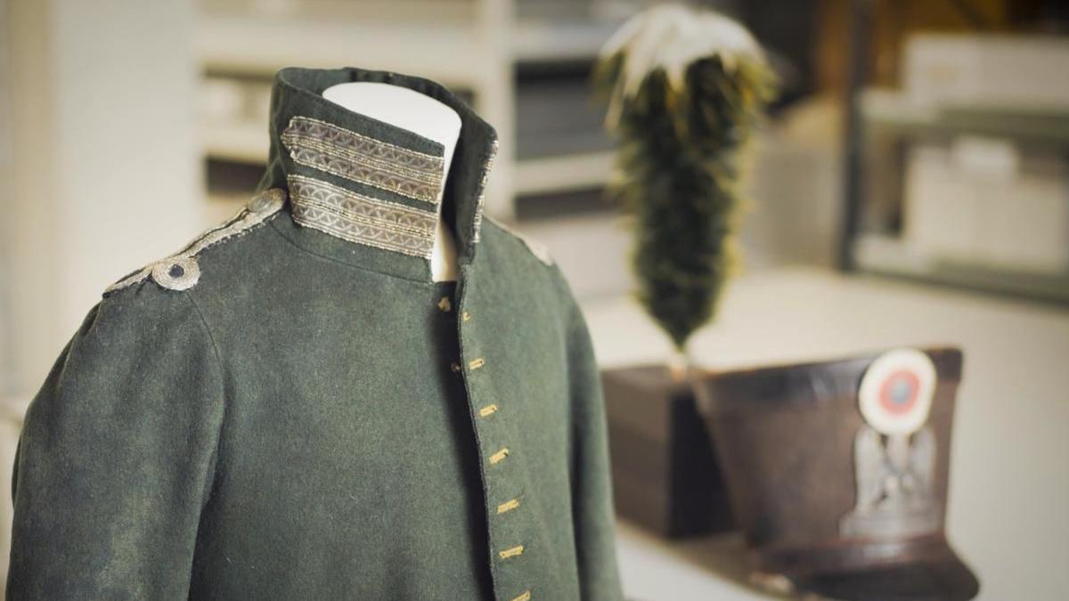 The army green jacket is worn and faded - this is a close up of the jacket, and the hat sits behind it, with the large green feather on its own base to the side.