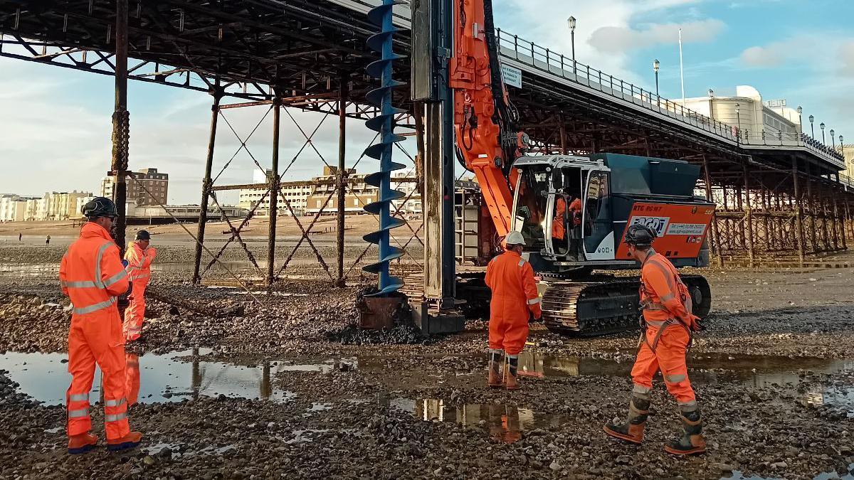 Four workers stood on the beach un Worthing, under the pier. They are all wearing orange high-vis outfits. Another man with a high-vis outfit is using a large industrial digger. 