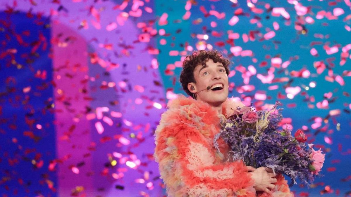 An elated-looking Nemo - the Swiss singer who won Eurovision in 2024. He has short, dark, curly hair, is wearing a coral pink fluffy coat and holding a large bunch of flowers. He is surrounded by falling sparkling confetti.