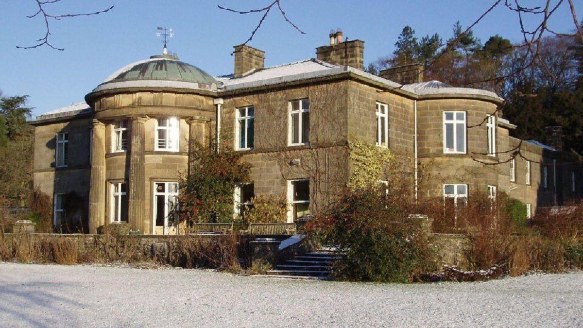 A large mansion-style building, pictured from the front. A frozen lawn covered in frost is in the foreground. A small set of steps lead up to the house itself, while there are also some shrubs around.