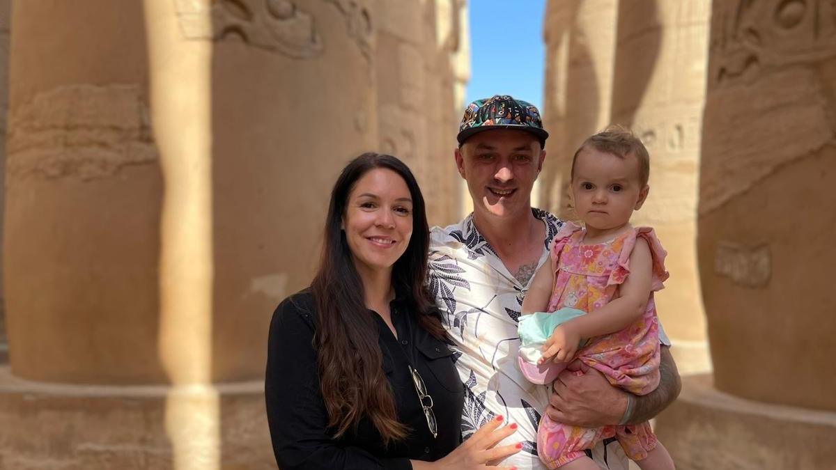 Paul and Ally standing between sandstone columns. Paul is holding their daughter who is wearing a pink dress. 