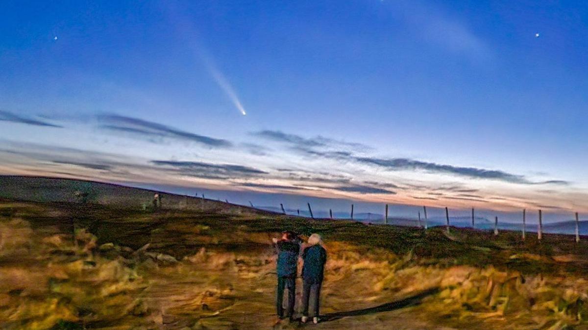 Two people looking at the comet. They are standing in a field and there are clouds and stars in the sky.