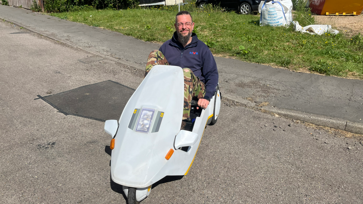 Mark Robinson is sitting in the vehicle. It is a triangular shape with three thin wheels, is light blue/grey in colour and has wing mirrors as well as a light on the front. Mr Robinson is smiling at the camera while holding onto the handle bars. The vehicle also has a tall flag on the back for other vehicles to see. 