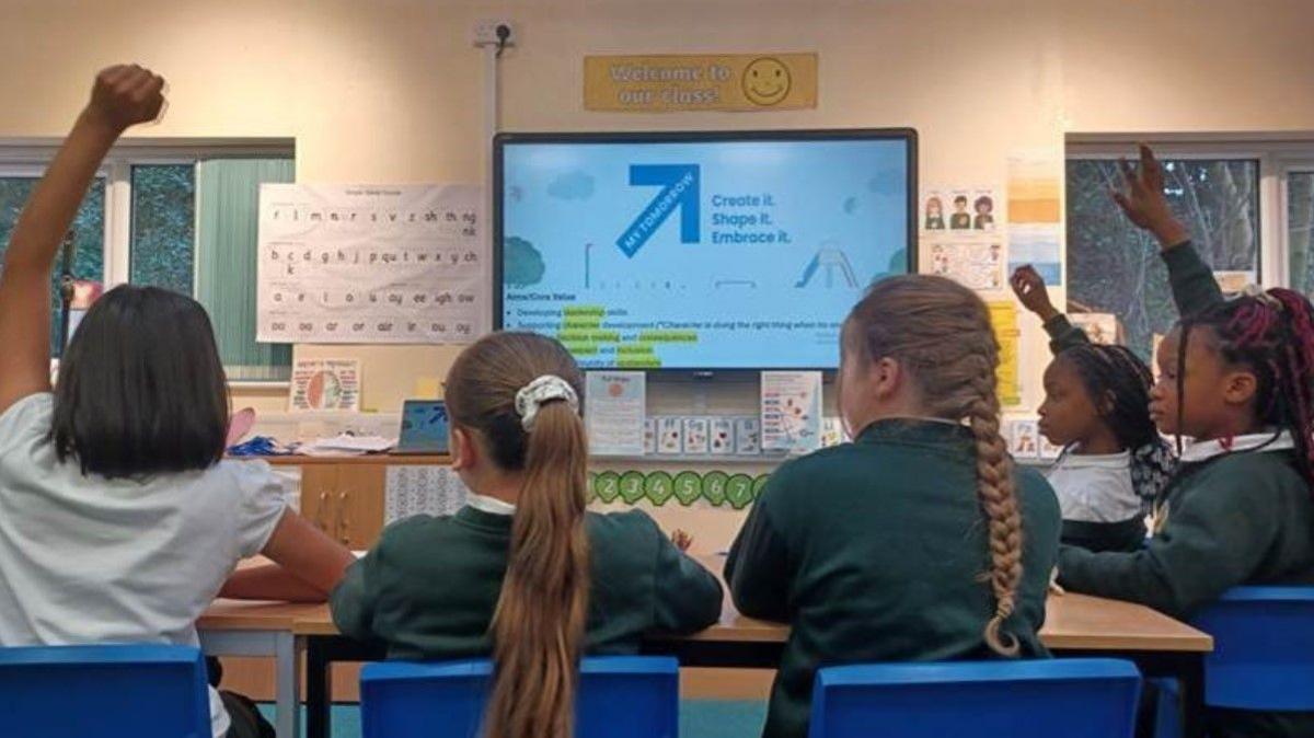 Five children in a classroom - three of them have their backs to the cameras and the others are side on to the camera. A screen is on a wall in the background, along with windows. 