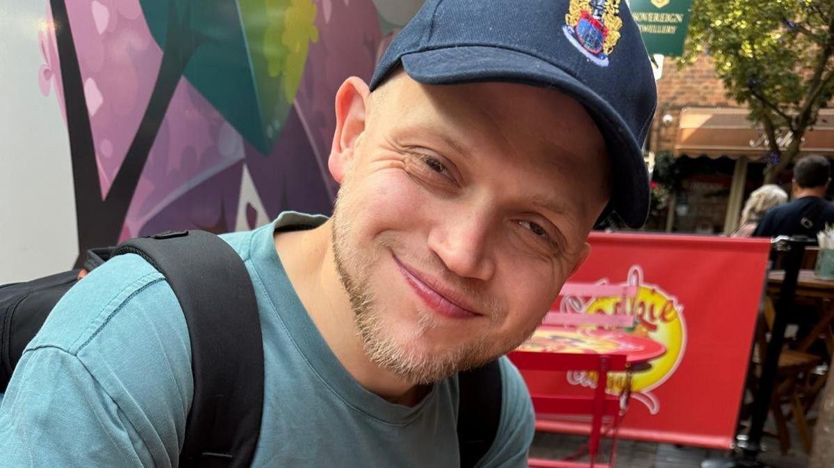 A man smiles at the camera wearing a baseball hat, blue T-shirt and black backpack. He is smiling at the camera and eating an ice cream. He is sat outdoors at an ice cream parlour