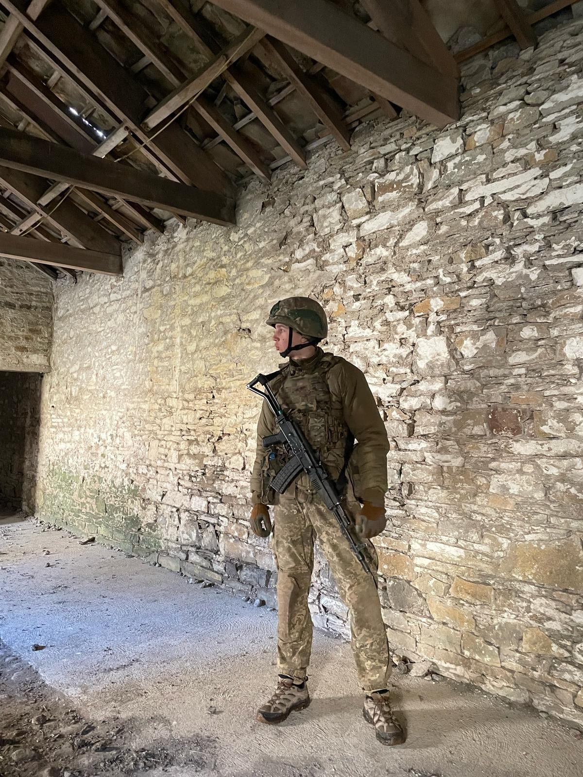 Maksym is dressed in military camouflage gear and has a rifle. He is standing against a stone wall.
