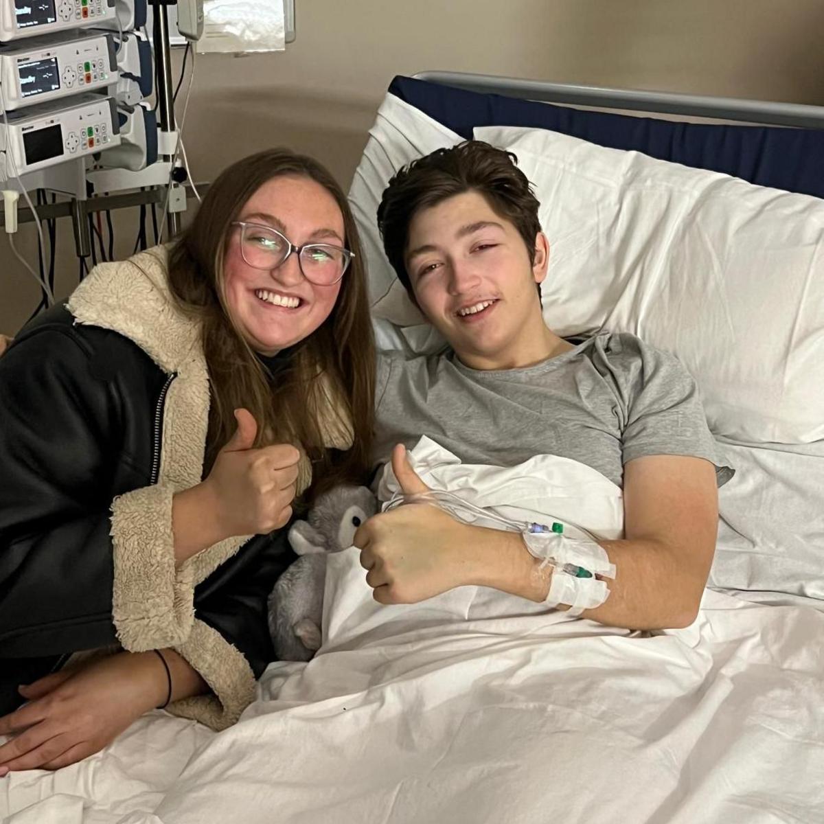 Jayme wearing a leather coat with a woolly trim around the neck with her thumb up next to Ted who is lying in his hospital bed with his thumb up too. Ted has IV lines attached to his arm