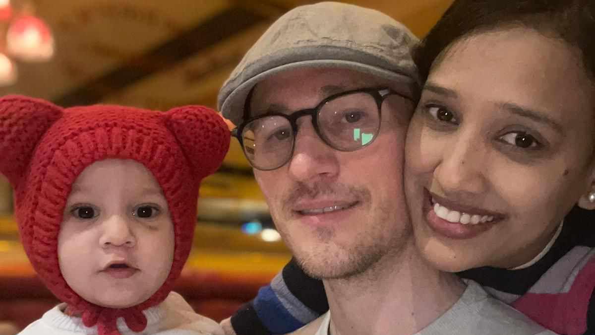 A small baby wearing a red, wrap-around woolly hat with ears, held next to Neha Dey-Morgan, who has black hair and is smiling with her cheek pressed against her husband's, who is in the middle of the photo. 