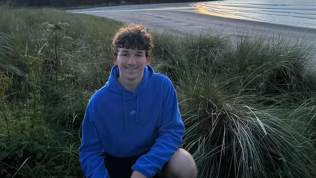 A teenager boy, with brown hair, kneeling in a patch of grass. There is a beach in the background. The boy is wearing black shorts and a blue hoodie. 