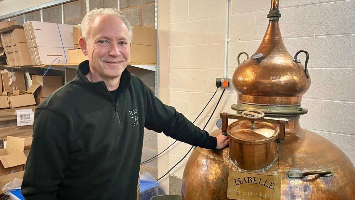 Mick Skerratt in a black fleece is standing with one hand on the copper distiller which is named Isabelle. There are numerous cardboard boxes in the background and two plugs connected to a power socket in the wall.