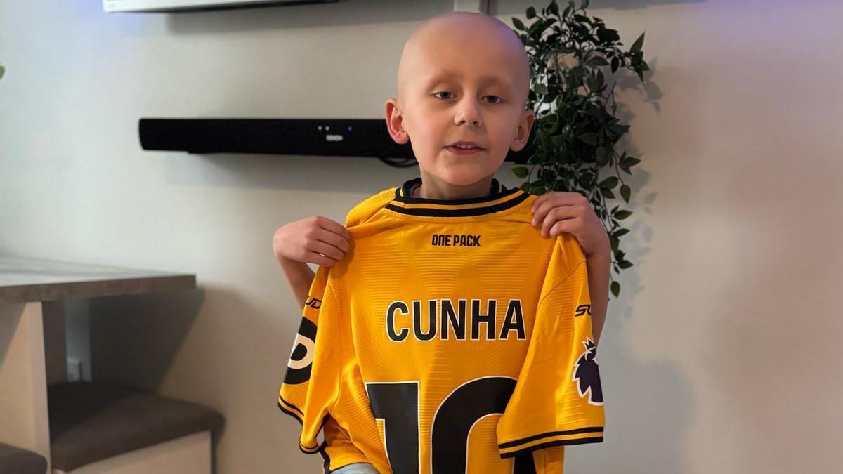 A young boy is pictured holding a yellow and black Wolverhampton Wanderers shirt with the name "Cunha" and the number ten on the back. He is standing in a toom with two shelves and a plant behind him, showing the shirt to the camera. He has no hair on his head due to cancer treatment.