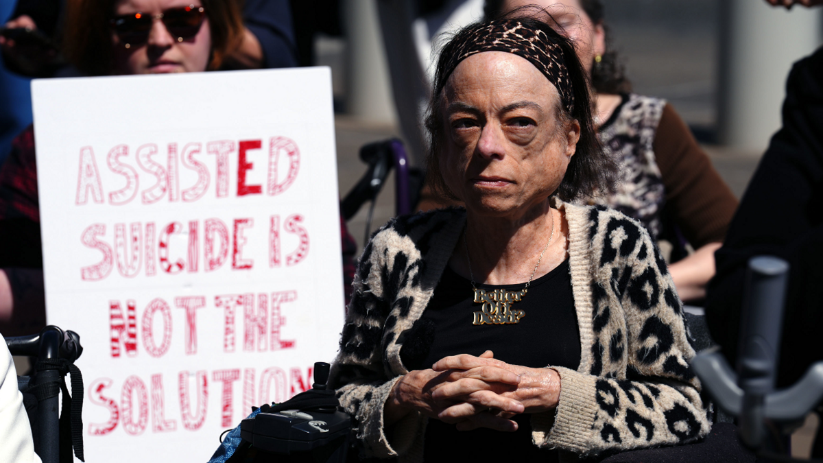 Actor Liz Carr, wearing a leopard print cardigan, black top and necklace. Next to her is a banner with the words 'Assisted suicide is not the solution'