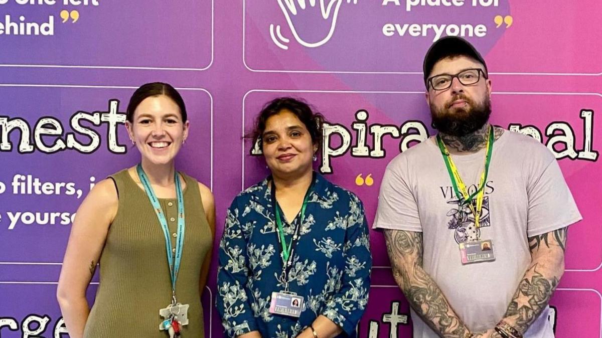 Two women and a man stand in front of a purple board with words on it and smile at the camera.