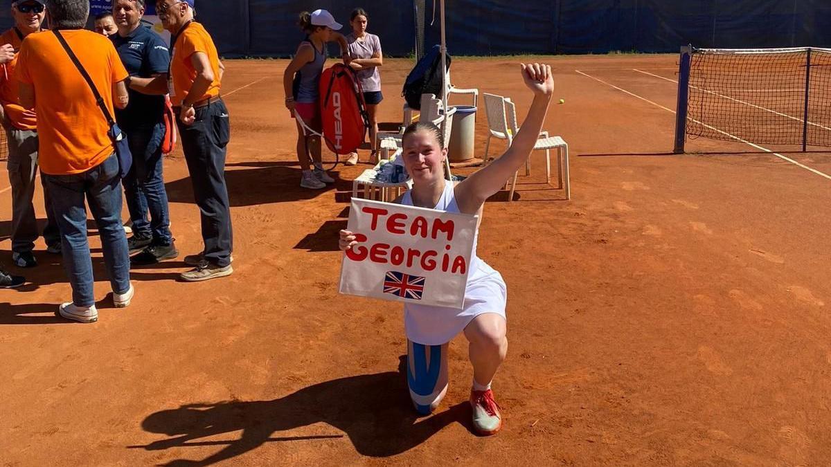 Georgia Routledge on one knee on the side of a clay tennis court, holding a 'Team Georgia' sign with a picture of the Great British flag.