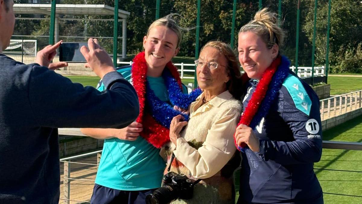 Hy Money, holding a camera, stands between two smiling women in sports gear, draped in red and blue garlands. A man takes their photo outdoors on a sports field.