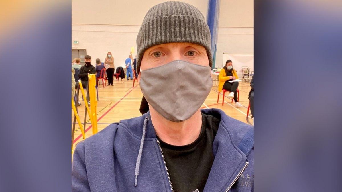 Larry wearing a grey hat and a grey facemask in a school sports hall which is dotted with people getting their covid jabs