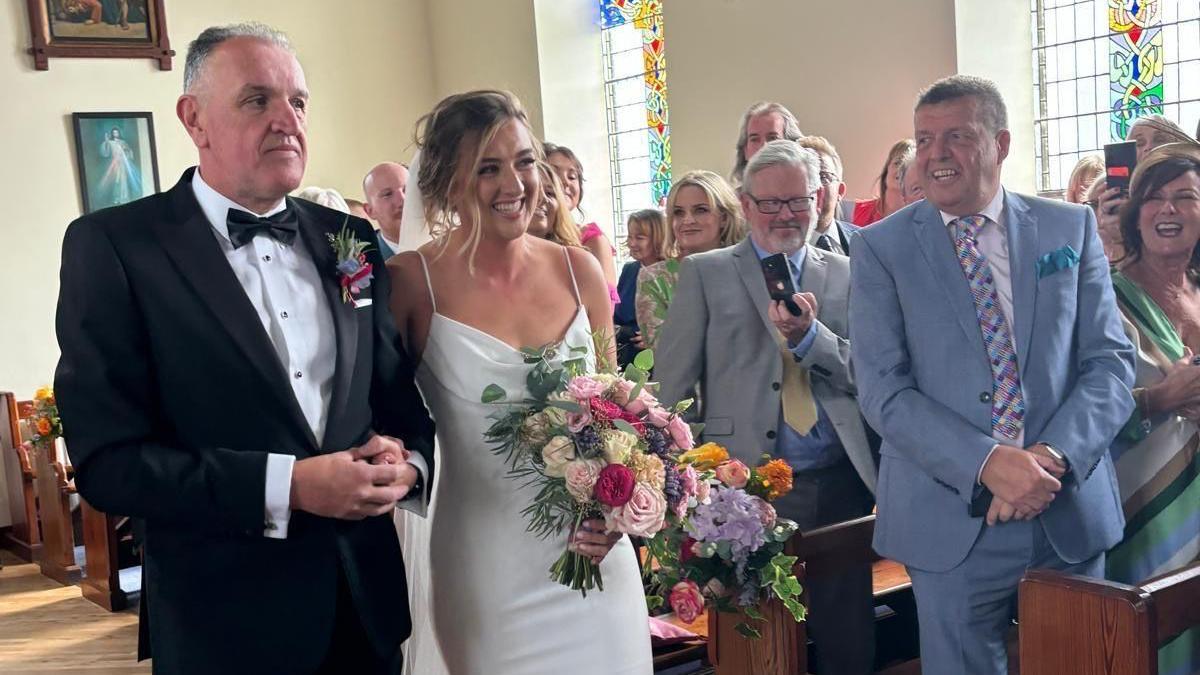 John in a black tuxedo walking his daughter down the aisle. She is smiling and is wearing a satin wedding dress with a veil trailing down her back from her hair. Her flowers are pink, red, white and purple. People are smiling in the pews to the right. 