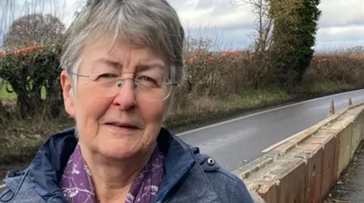 A woman with grey hair and a blue coat standing by a rural road with a hedge running alongside it.