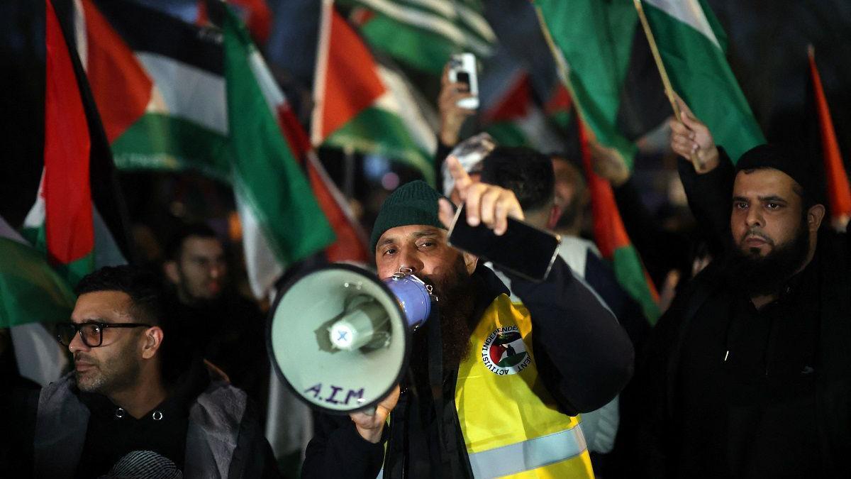 Pro-Palestinian protesters gather outside the stadium before the match. A man with a hi-vis jacket has a loudspeaker. The group are carrying flags.
