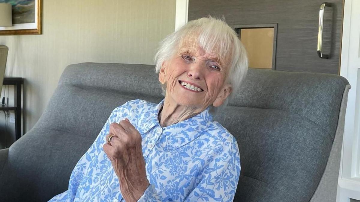 An elderly woman with white hair, wearing a pale blue blouse with a darker blue flower pattern. She is sitting on a small grey and smiling as she looks at something beyond the camera. 