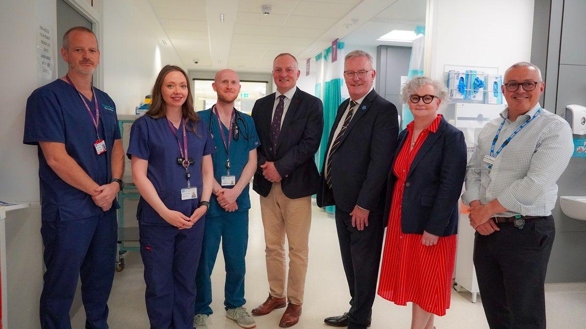Mike Nesbitt has grey hair and glasses and is wearing a dark suit while standing among hospital staff, some who are in blue scrubs. They are standing on a hospital ward.