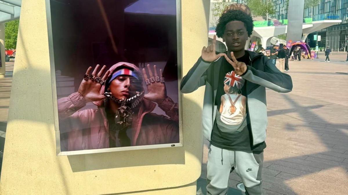 Carlos poses in front of a poster for rapper Central Cee, who recently collaborated with Drake. Carlos is wearing a grey tracksuit and t-shirt, and has black hair.