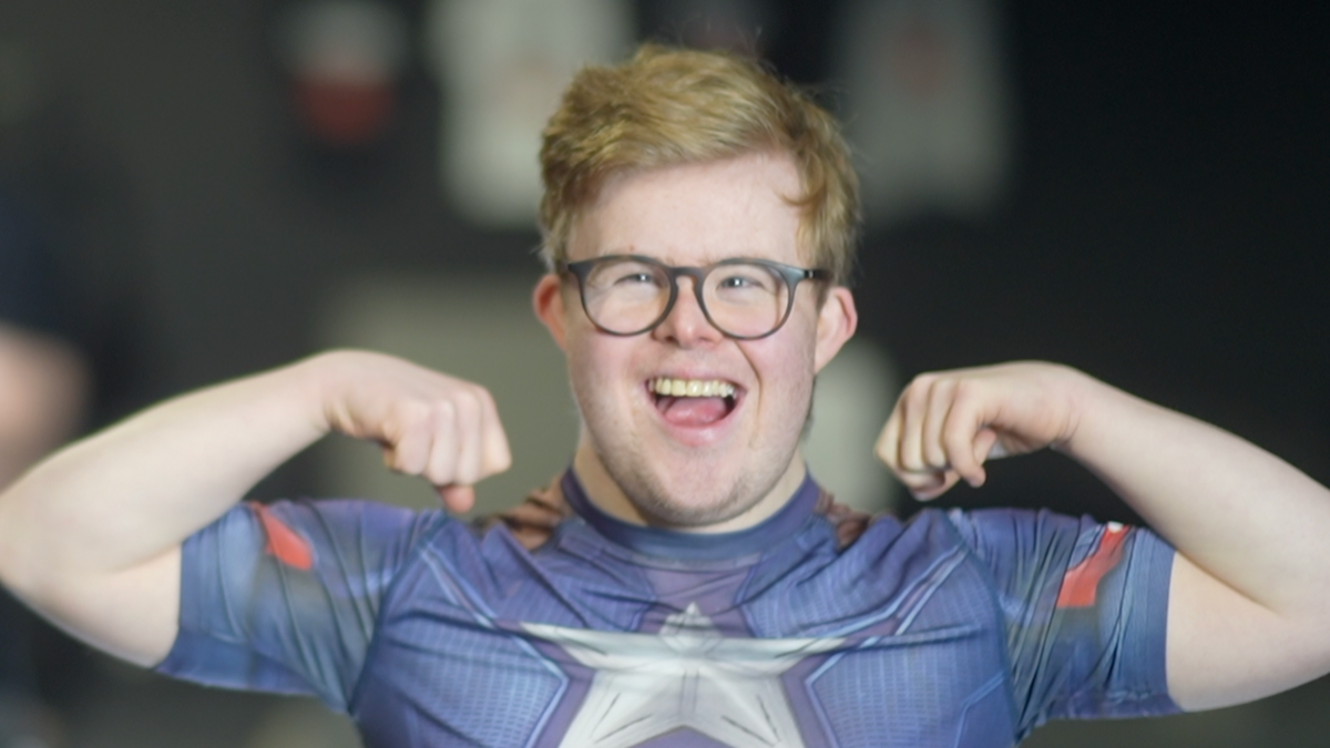 Edward Wagland with blonde hair smiles widely at the camera while tensing his bicep muscles in his arms. He is wearing a blue top with a silver star in the middle of it as well as black glasses.