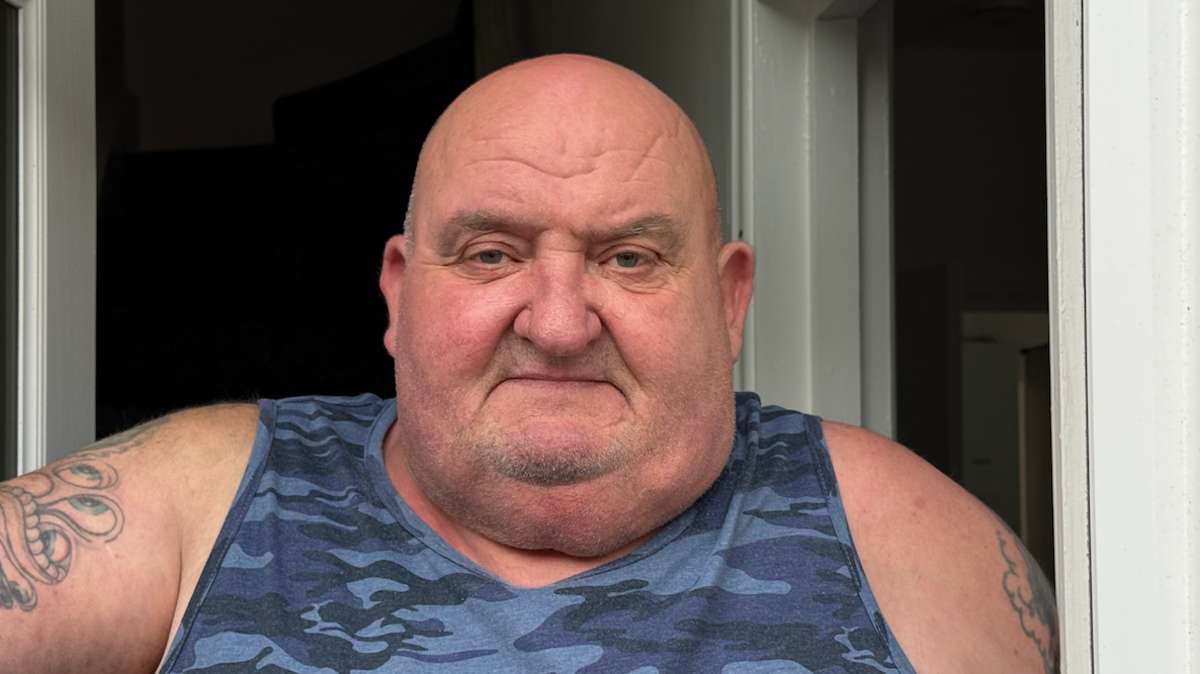 Graham Horn, who is wearing a blue coloured camouflaged vest and with tattoos on both arms, stands in his doorway.