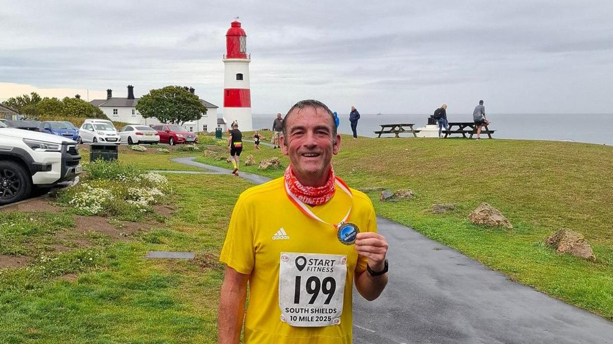 John Paul Green at Souter Lighthouse in Whitburn. He is wearing a yellow running vest and holding a medal in his left hand having completed the South Shields 10 Mile race. The lighthouse is painted red and white.