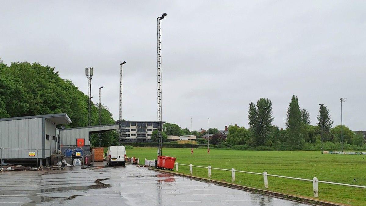 A rugby pitch with car park in the foreground. There is a block of flats in the distance, overlooking the club, and a club house and spectator stand to the left hand side of the image.