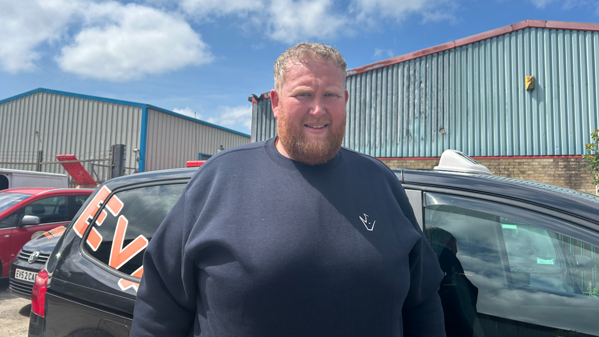 Evan Shepherd mewn siwmper ddu o flaen tacsi