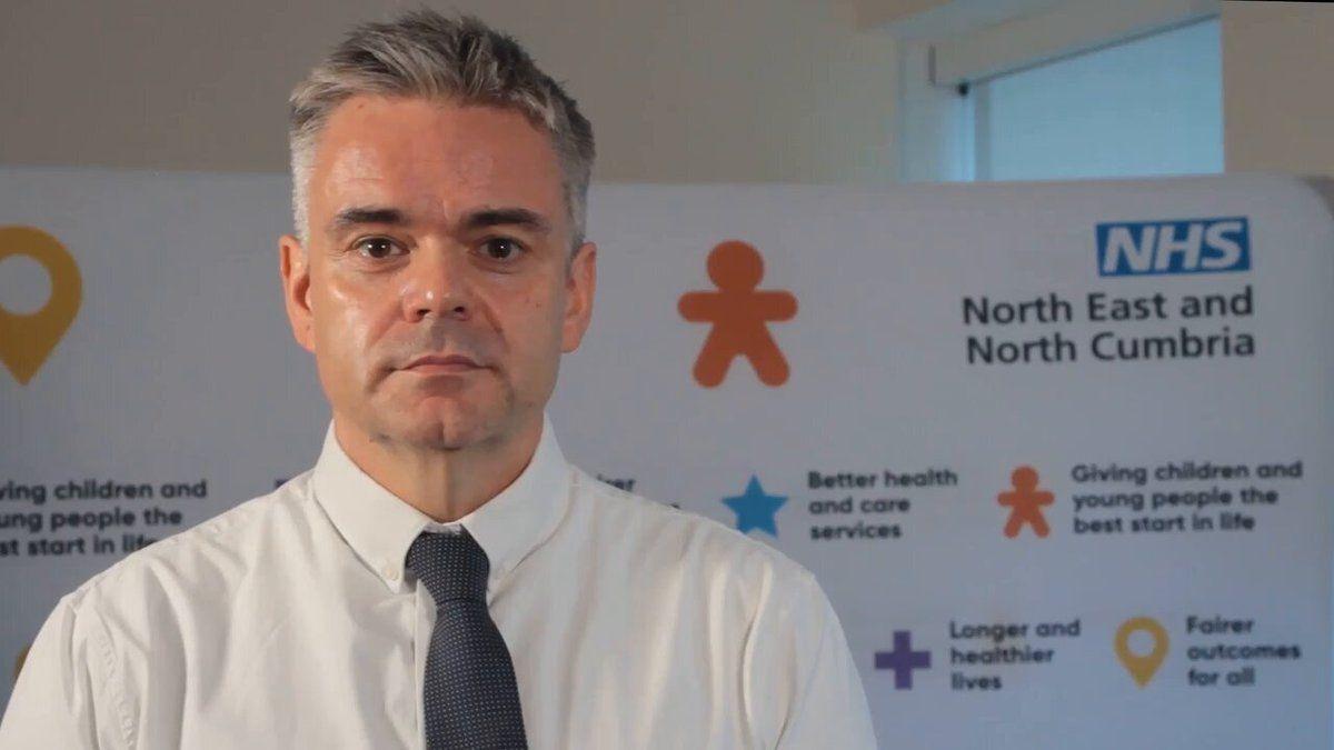 Dr Neil O'Brien standing in front of a large white board containing, in the top corner, the NHS logo above the words North East and North Cumbria. There are a number of symbols including a purple cross, a blue star, the shape of a person coloured orange and a location symbol along with a series of marketing-style phrase such as "Better health and care services" and "Fairer outcomes for all". Dr O'Brien has short, neat silvery grey hair and is wearing a dark grey tie with small white dots on it and a smart white shirt. He has a dimple in the centre of his chin, thick black eyebrows and dark eyes.