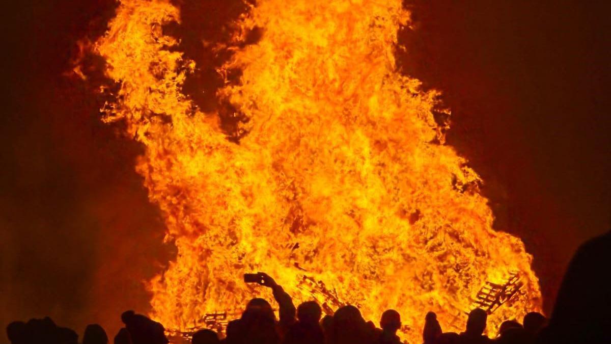 A large fire burning with small silhouettes of people in front of it, one taken a photo on their phone.