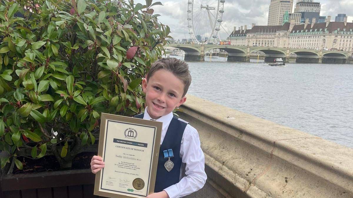 Teddy a young boy wearing a shirt and waistcoat with a medal pinned to it he is holding a certificate with the London Eye and River Thames behind.