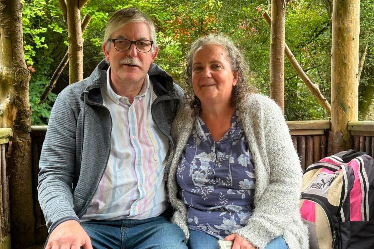 A man and a woman in their 60s sit in an open sided tree house where the trunks of the trees behind them are shown to hold up the platform. There is foliage in the background. The couple are sitting close together and are both wearing jeans. The man has glasses and a pastel striped shirt and is wearing an unzipped grey fleece over it. The woman has a blue top with white flowers with a grey fluffy cardigan over it. There is a black, pink and beige rucksack next to her.