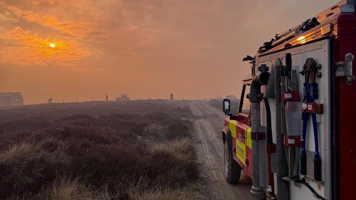 A fire engine on a moor which is bathed in orange light, part from the sunset and part from the light of the fire. 