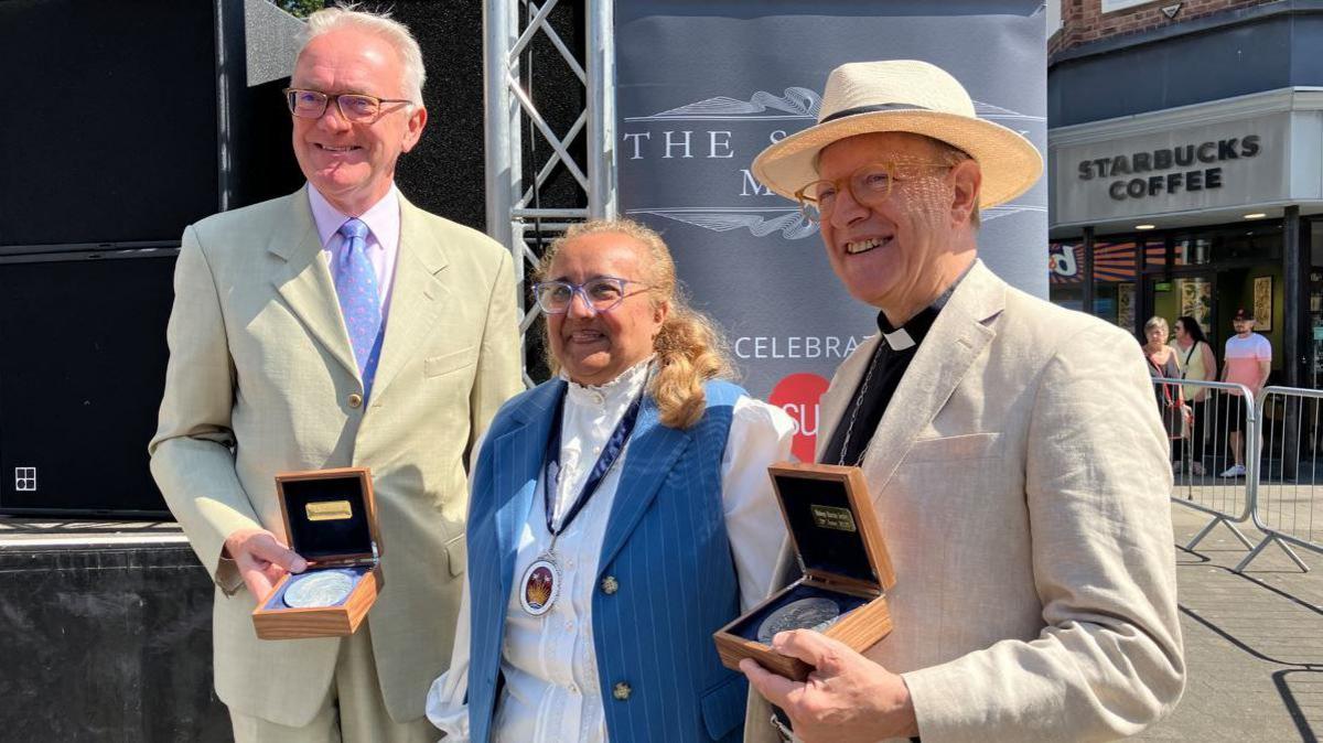 Suffolk High Sheriff Gulshan Kayembe, pictured centre, with medal recipients Martin Seeley and Mark Pendlington. The two men are wearing white suits and holding their medals. Gulshan Kayembe is wearing a white shirt and blue waistcoat. They are pictured in Lowestoft town centre, with a coffee shop in the background