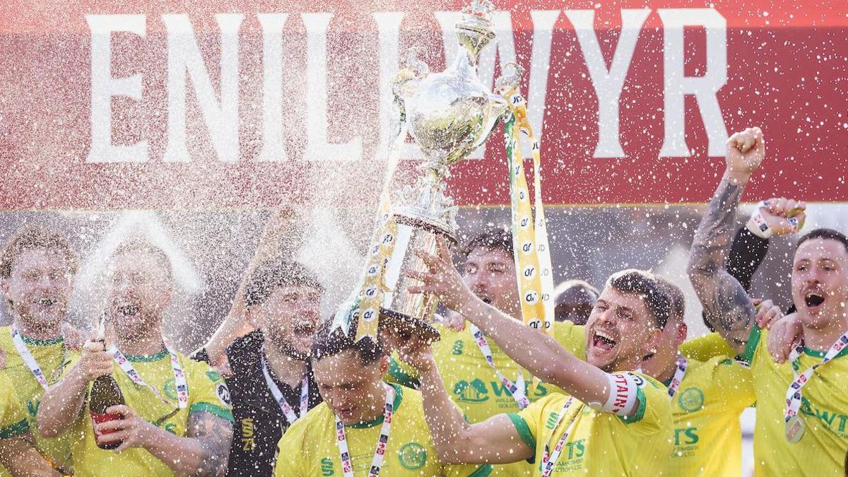 Caernarfon players in yellow spray champagne and hold the trophy as they celebrate their Welsh Cup final win
